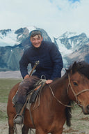 A Mongolian herder on horseback smiling at the camera, snowy peaks behind