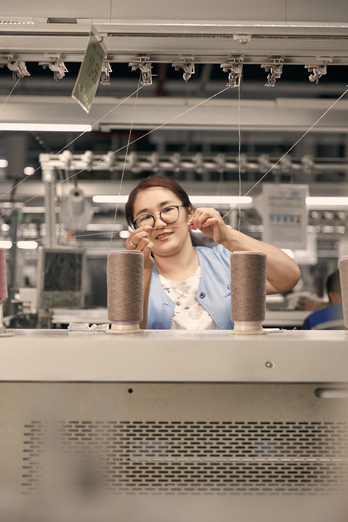 A GOBI knitting technician in a blue vest threading yarn at a knitting machine, cones of cashmere in front of her