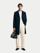 Man wearing a navy coat, white shirt, beige pants, and holding a brown leather bag on a white background