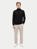 A man modeling a black long-sleeve turtleneck sweater, paired with beige trousers and white sneakers, against a light gray studio background.