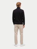 A man viewed from the back wearing a black mock-neck knit sweater, beige trousers, and white sneakers, standing against a light gray studio background.