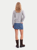 A woman is shown from behind wearing a light gray long-sleeve sweater, a blue denim mini skirt, and dark brown boots, standing against a solid off-white background.