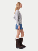 A woman in side profile wearing a light gray long-sleeve knit sweater, a blue denim mini skirt, white socks, and dark brown mid-calf boots, photographed against a plain light gray studio background.