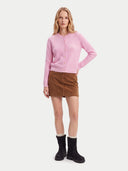 A woman modeling a light pink cable-knit cardigan sweater, brown mini skirt, and black ankle boots against a light gray studio background.