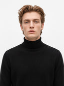 A man with light brown hair wearing a black ribbed turtleneck sweater, photographed against a clean light gray background.