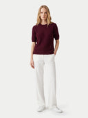 A woman stands against a light gray studio background wearing a short-sleeved ribbed knit sweater in deep burgundy, paired with white wide-leg trousers and white sneakers.