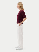 A woman stands in profile wearing a short-sleeve ribbed burgundy top, white trousers, and white sneakers against a plain light gray studio background.