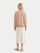 A woman viewed from the back wearing a beige knit mock-neck sweater and a cream midi skirt, paired with beige flat shoes, against a light gray studio background.