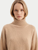 A woman with a short blonde bob wearing a beige ribbed turtleneck sweater, photographed against a clean light gray studio background.