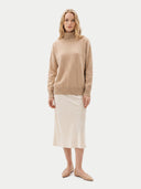 A woman modeling a beige turtleneck sweater and a light cream midi skirt, paired with beige pointed-toe flats, against a solid light gray background.