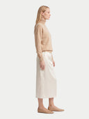 A woman stands in profile wearing a beige turtleneck sweater, a cream midi skirt, and beige flats against a plain light gray studio background.
