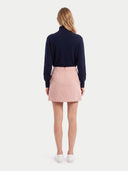 A woman viewed from the back wearing a navy blue mock-neck long-sleeve top and a light pink mini skirt, paired with white sneakers, against a light gray studio background.