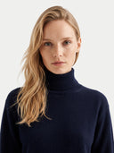 A blonde woman with freckles wearing a dark navy blue ribbed turtleneck sweater, photographed against a clean light gray background.