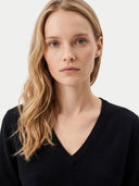 A close-up portrait of a woman with blonde hair wearing a black V-neck knit sweater against a light gray background.