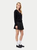 A blonde model stands in profile wearing a black long-sleeve V-neck sweater, a black pleated mini skirt, white socks, and black loafers against a light gray studio background.