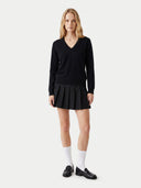 A blonde model stands against a light gray studio background wearing a black long-sleeve V-neck sweater, a black pleated mini skirt, white knee-high socks, and black loafers.