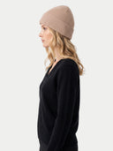 A woman in profile wearing a ribbed knit beanie in a dusty rose-beige color and a black V-neck sweater, photographed against a clean light gray studio background.