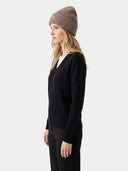 A woman in profile wearing a ribbed taupe beanie, a black long-sleeve V-neck sweater, and black trousers, photographed against a light gray studio background.