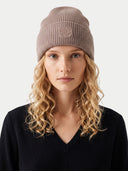A woman with long curly blonde hair wearing a ribbed knit beanie in taupe and a black V-neck sweater, photographed against a light gray studio background.