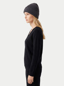 A woman in side profile wearing a dark charcoal ribbed knit beanie, a black long-sleeve V-neck sweater, and black pants, photographed against a light gray studio background.