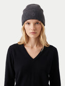 A woman wearing a dark charcoal gray ribbed knit beanie with a subtle embroidered logo on the front, paired with a black V-neck sweater, against a light gray studio background.