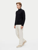 A man wearing a black cable-knit mock-neck sweater, cream-colored trousers, and beige sneakers, standing against a plain light gray studio background.