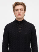 A man wearing a black cable-knit mock-neck sweater with a three-button placket, photographed against a plain light gray studio background.