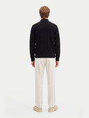 A man viewed from the back wearing a black cable-knit sweater, cream trousers, and beige shoes, standing against a light gray studio background.