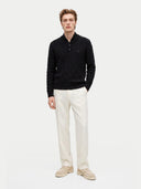A male model wearing a black cable-knit polo sweater, off-white trousers, and beige loafers, standing against a plain light gray studio background.