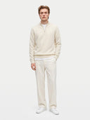 A man modeling a cream-colored knit half-zip sweater, matching cream trousers, and white sneakers against a solid light gray background.