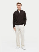 A man modeling a dark brown quarter-zip knit sweater over a white undershirt, paired with off-white trousers and white sneakers, against a plain light gray studio background.
