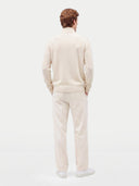 A man viewed from behind wearing a cream-colored knit sweater and matching trousers, paired with white sneakers, against a plain light gray studio background.