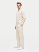 A man modeling a matching cream-colored knit set consisting of a half-zip mock neck sweater and wide-leg trousers, paired with white sneakers, against a solid light gray background.