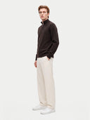 A man modeling a dark brown quarter-zip mock-neck sweater, cream wide-leg trousers, and white sneakers against a light gray studio background.