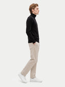 A man in profile wearing a black turtleneck sweater, beige trousers, and white sneakers, standing against a light gray studio background.