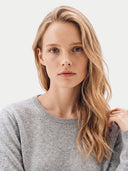 A blonde woman with freckles wearing a light gray crewneck sweater, shown in a close-up portrait against a clean white background.