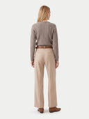 A woman is shown from the back wearing a taupe cable-knit sweater, beige wide-leg trousers, a brown belt, and brown leather shoes against a light gray studio background.