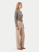 A woman stands in profile wearing a taupe cable-knit sweater, beige wide-leg trousers with a brown belt, and brown shoes against a plain light gray studio background.