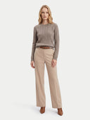 A woman stands against a light gray studio background wearing a taupe cable-knit sweater, beige wide-leg trousers, a brown belt, and brown ankle boots.