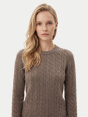 A blonde woman wearing a brown cable-knit crewneck sweater, photographed against a clean light gray studio background.