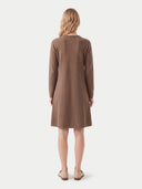 A woman viewed from the back wearing a brown long-sleeve knit dress, standing against a plain light gray studio background.