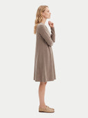 A woman stands in profile wearing a long-sleeve, knee-length knit dress in a taupe-brown color, paired with beige loafers, against a solid off-white background.
