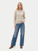A woman modeling a light beige mock-neck knit sweater and wide-leg blue jeans with brown pointed-toe boots, standing against a plain light gray studio background.