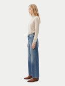 A woman stands in profile wearing a light beige long-sleeve knit top, high-waisted wide-leg blue jeans, and brown ankle boots against a plain light gray studio background.