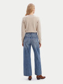 A woman viewed from the back wearing a light beige knit sweater, wide-leg blue jeans, and brown leather boots against a plain light gray studio background.
