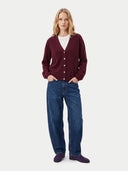 A woman stands against a light gray studio background wearing a burgundy button-front cardigan over a white top, blue jeans, and dark slip-on shoes.
