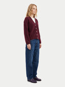 A woman stands in profile wearing a burgundy button-front cardigan over a white top, blue jeans, and dark loafers against a plain light gray studio background.
