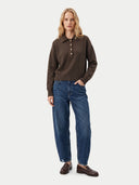 A woman stands against a light gray studio background wearing a dark brown long-sleeve polo sweater with a button placket, blue jeans, and brown leather loafers.
