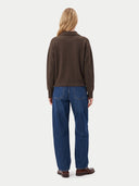 back view of a woman standing wearing a dark brown sweater with a collar and blue jeans on a light gray background.
