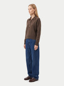 A woman stands in profile wearing a brown long-sleeve polo sweater, wide-leg blue jeans, and brown leather shoes against a light gray studio background.